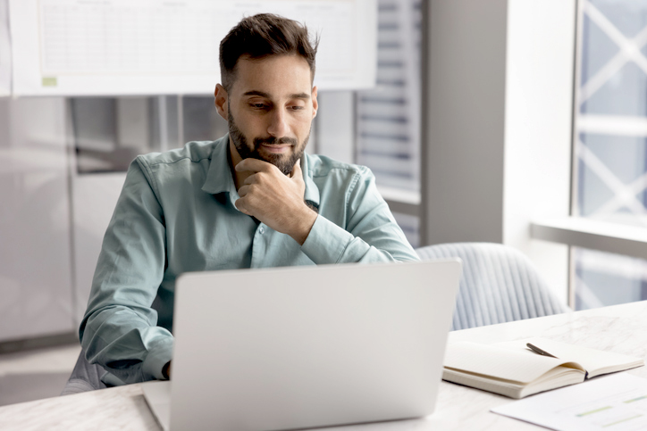 Focused Latin businessman, office employee sits at desk look at laptop screen, watching on-line training or professional webinar, reviewing business presentation, researching information for project