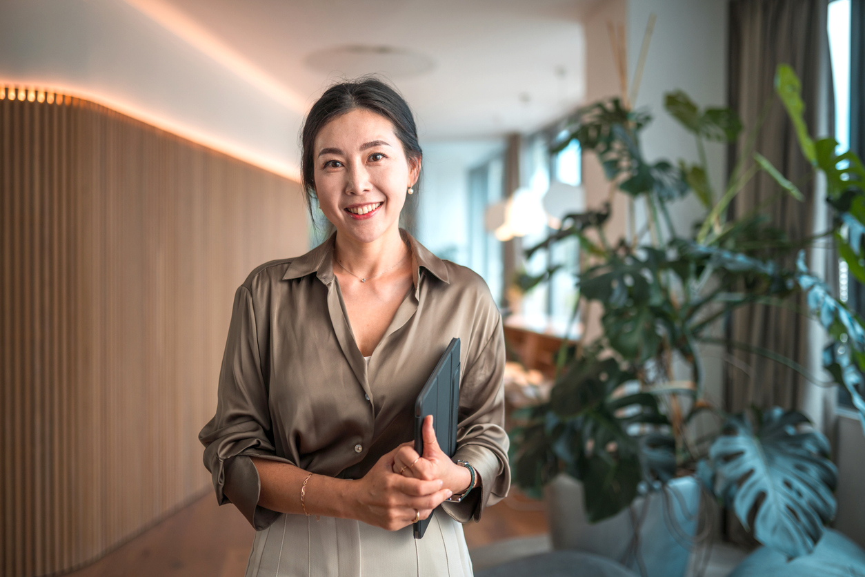 Positioned within a modern corporate environment, a mid-aged Asian businesswoman of striking beauty stands in her office, poised with a digital tablet, using it to execute digital tools and wireless technology to perfection in her strategic endeavors.