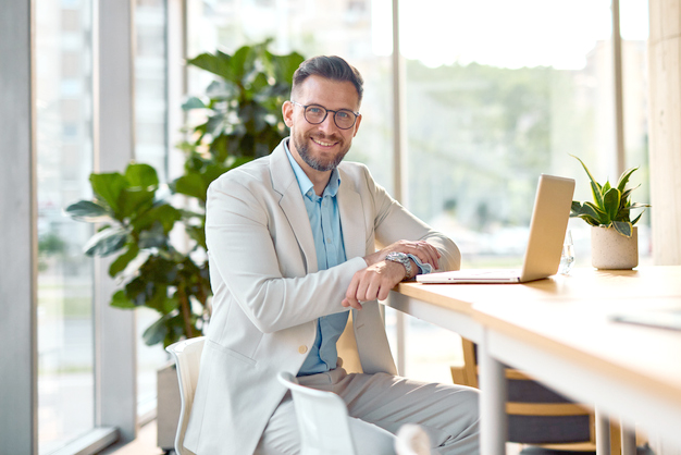 Cheerful businessman sitting at a desk and working on a laptop in a modern office, exuding confidence while smiling and looking at the camera, surrounded by a bright, inviting interior