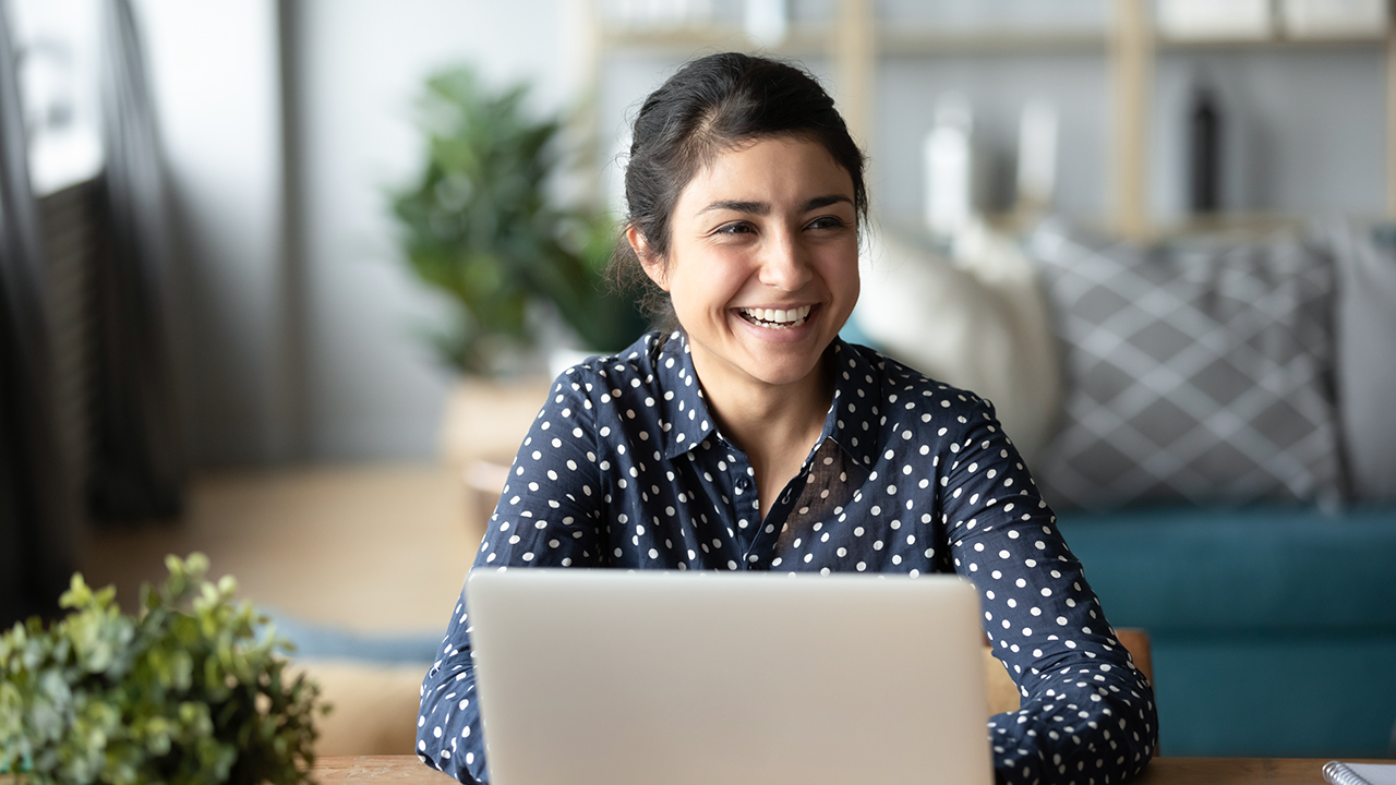 Happy cheerful millennial indian business woman sitting at desk in front of laptop laughing talking to someone in home office room looking into distance feels overjoyed. Successful freelancer portrait